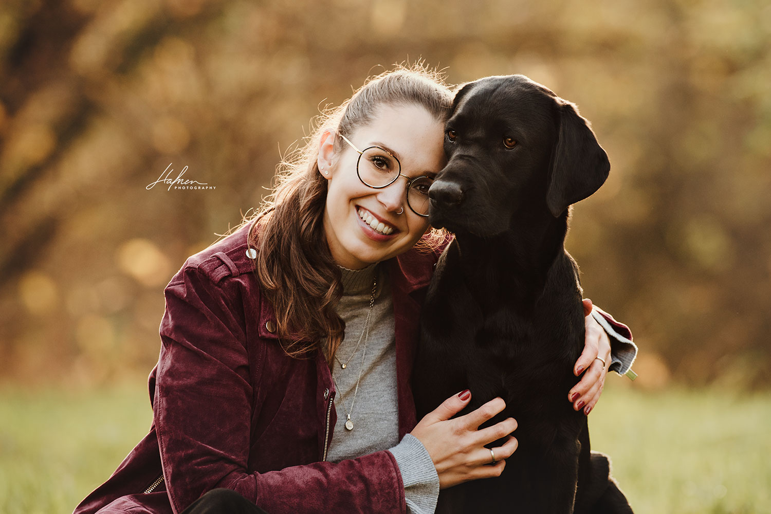 Eine lächelnde Frau in einer roten Jacke sitzt neben einem schwarzen Hund, beide schauen in die Kamera.