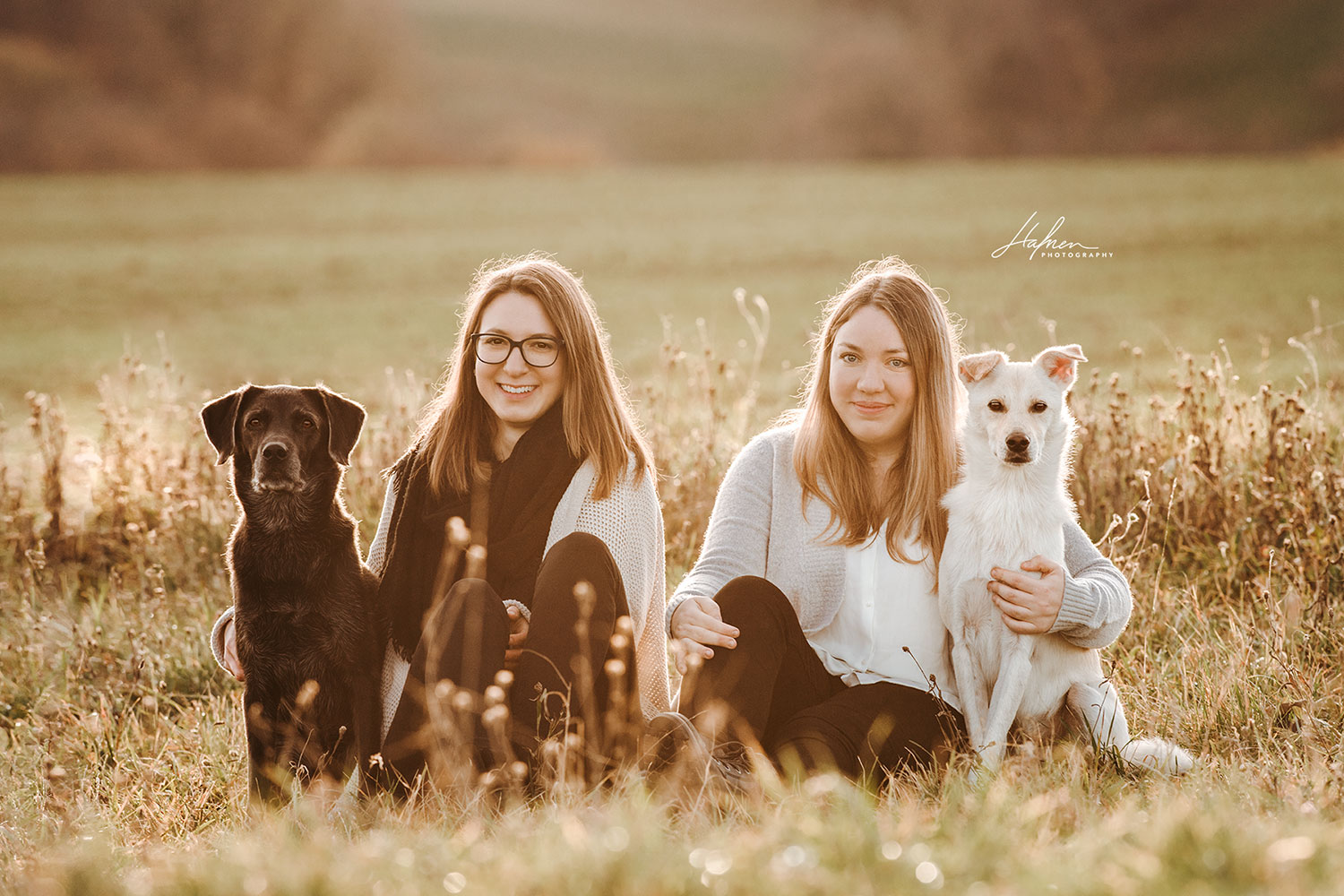 Zwei Frauen sitzen auf einer Wiese, jede hält einen Hund, eine Frau trägt eine Brille und einen schwarzen Schal, die andere Frau hat ein weißes Hemd und eine graue Strickjacke an.