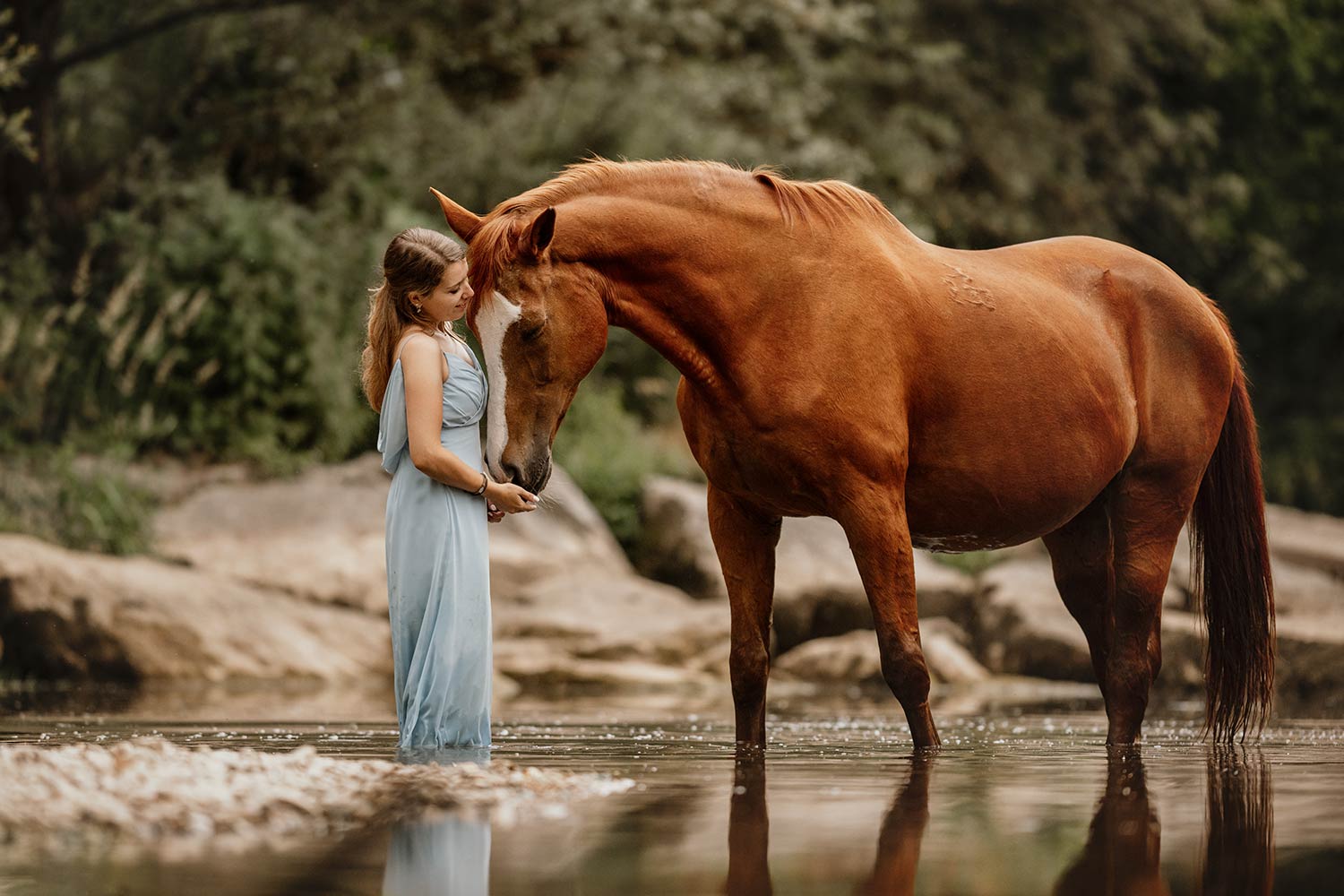Eine junge Frau in einem hellblauen Kleid steht in einem Fluss und berührt zärtlich die Nase eines fuchfarbenen Pferdes, das sich liebevoll zu ihr neigt.