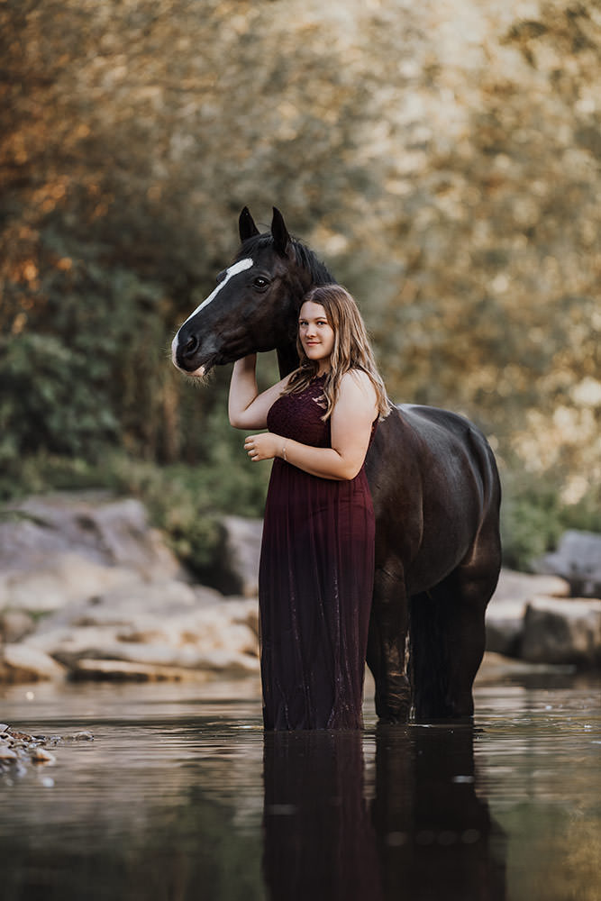 Eine Frau im burgunderfarbenen Kleid steht im Wasser und hält liebevoll ein schwarzes Pferd umschlungen.
