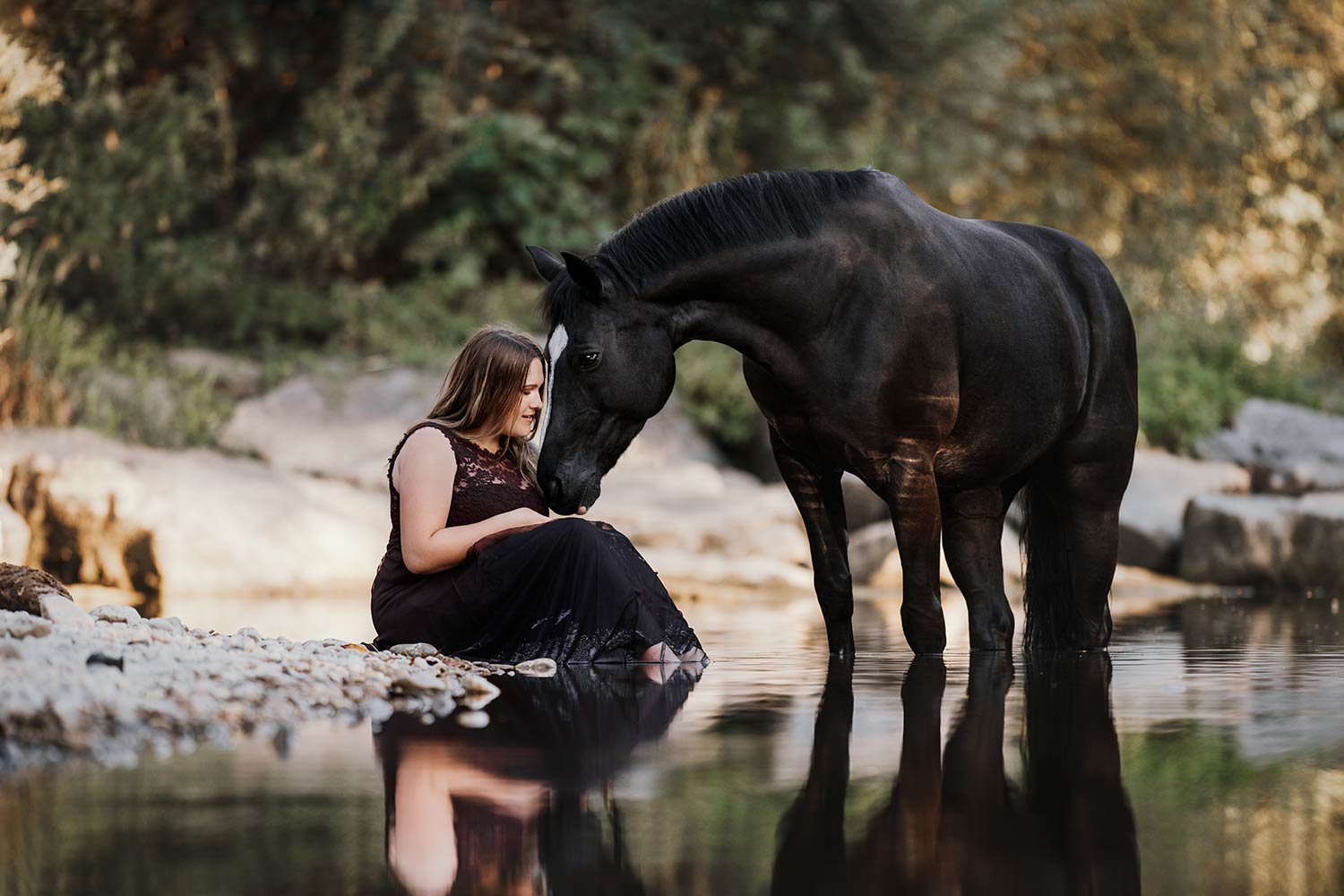 Eine Frau in einem dunklen Kleid sitzt am Ufer eines stillen Gewässers und neigt sich einem grossen schwarzen Pferd zu, das den Kopf sanft zu ihr neigt.