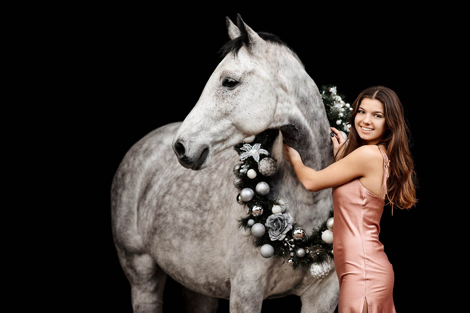 Eine lächelnde junge Frau in einem rosafarbenen Kleid schmückt ein apfelschimmel Pferd mit einem Weihnachtskranz.
