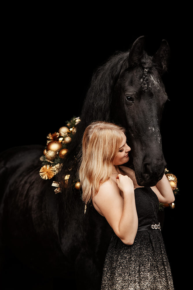 Eine blonde Frau in einem funkelnden Kleid umarmt liebevoll ein schwarzes Pferd, das mit einem goldfarbenen Weihnachtskranz geschmückt ist.