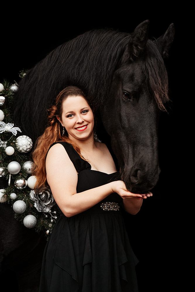 Eine lachende Frau in einem schwarzen Kleid steht neben einem Friesen mit einem weissen Weihnachtskranz um den Hals.