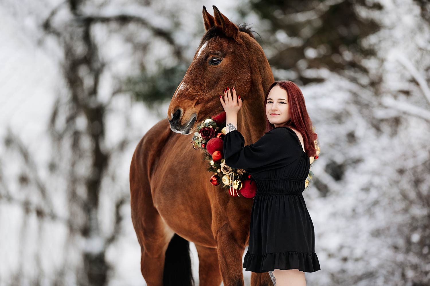 Eine Frau mit roten Haaren umarmt liebevoll ein braunes Pferd, das einen Weihnachtskranz aus roten Kugeln trägt.