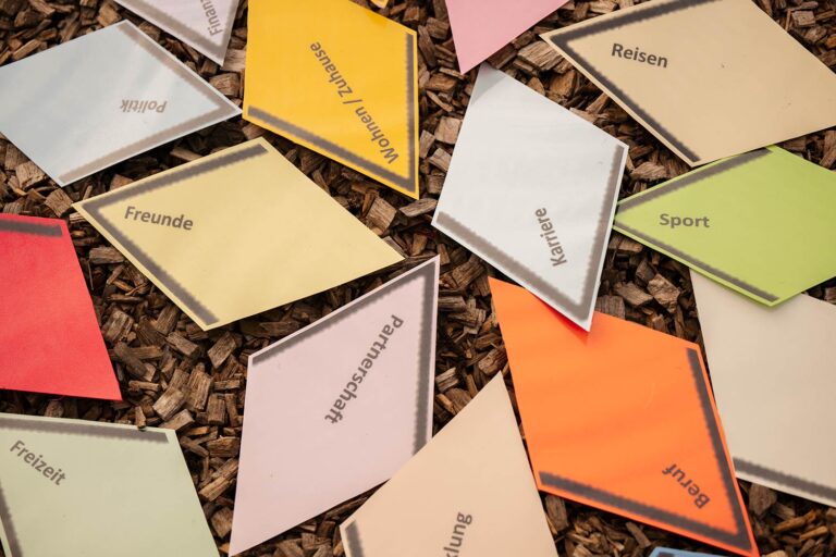 Farbige Zettel mit verschiedenen Lebensbereichen wie „Freunde“, „Karriere“ und „Sport“ liegen auf Holzhackschnitzeln.
