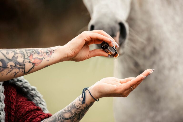 Eine tätowierte Person hält eine Flasche ätherisches Öl, während ein Pferd neugierig zusieht - professionelle Pferdefotografie