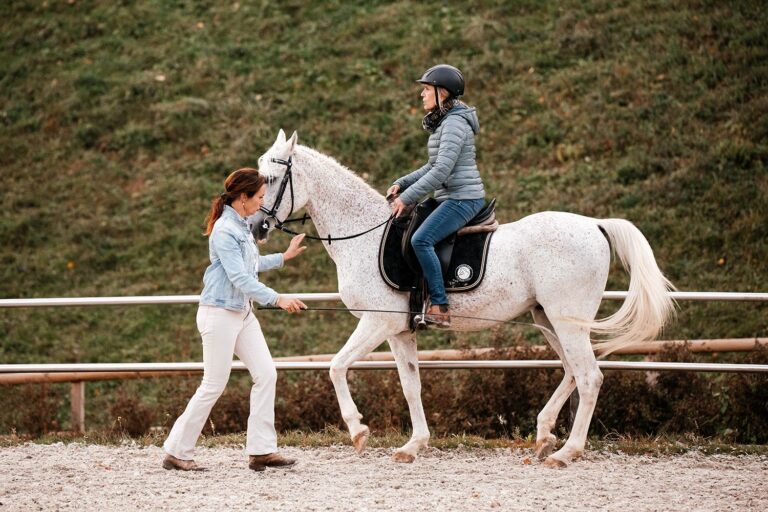 Eine Reitlehrerin gibt Anweisungen an eine Reiterin auf einem weissen Pferd in einer Reitarena.