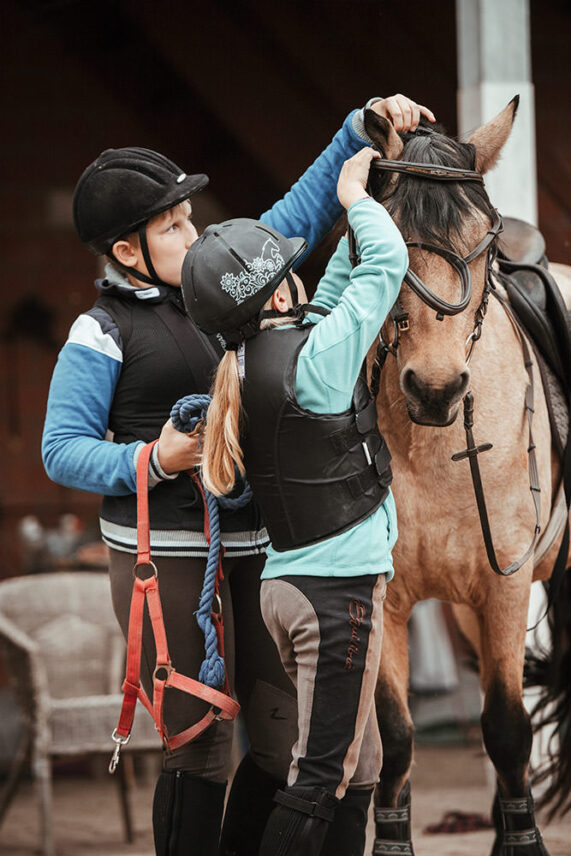 Zwei Kinder in Reitausrüstung helfen sich gegenseitig, das Zaumzeug auf ein Pony zu setzen.