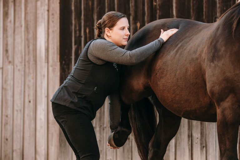 Eine Frau führt Dehnübungen mit einem braunen Pferd vor einer Holzwand durch.