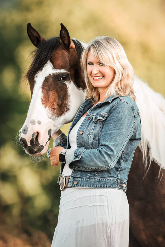 Eine lächelnde Frau in einer Jeansjacke steht neben ihrem gescheckten Pferd in einer malerischen Landschaft.