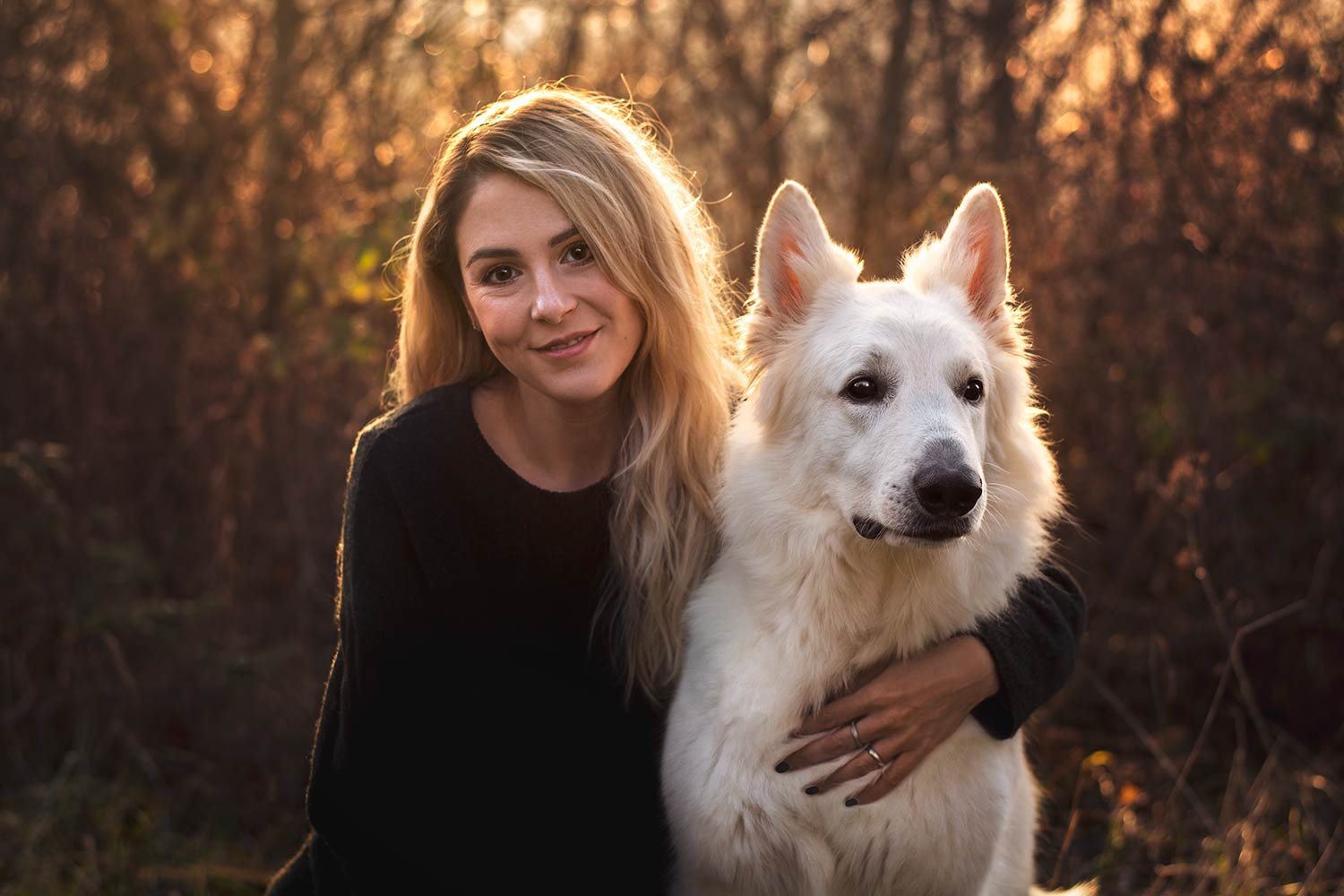 Eine junge Frau in einem schwarzen Pulli hält liebevoll einen weissen Hund im Arm und beide schauen in die Kamera.