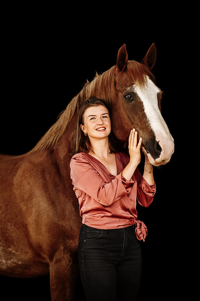 Eine Frau umarmt liebevoll ein braunes Freiberger Pferd vor schwarzem Hintergrund.